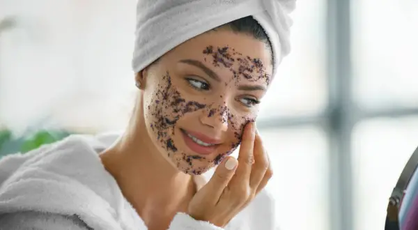 Femme souriante appliquant un gommage sur son visage. Elle porte une serviette sur la tête et un peignoir. La question est : combien de temps laisser un gommage sur le corps ?