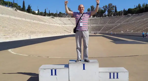Mężczyzna na podium w historycznym stadionie panatenajskim, z rękami w górze, świętuje zwycięstwo.