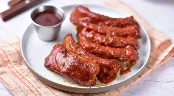 Soczyste żeberka w sosie BBQ, podane na talerzu z małą miseczką sosu. Idealne na letniego grilla.