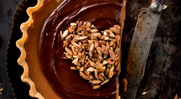 Herzförmige Schoko Tarte mit Karamell und gehackten Mandeln, daneben ein altes Messer.