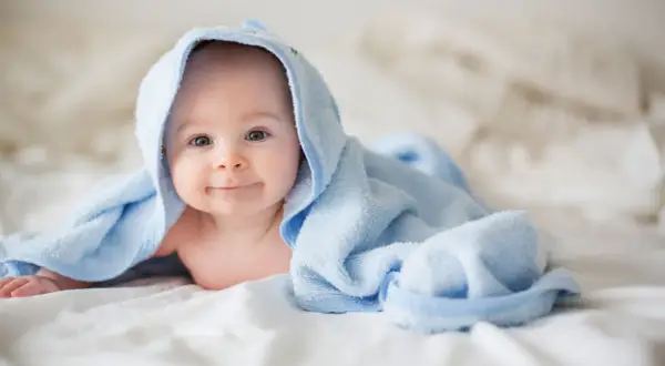 Ein lächelndes neugeborenes Baby liegt auf einem weißen Bett, eingewickelt in ein hellblaues Handtuch.
