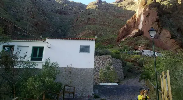 Un niño toma fotos en un camino empedrado, con una casa blanca y montañas rojas de fondo, cerca de la cueva bermeja.