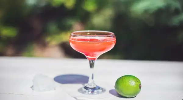 Orzeźwiający strawberry daiquiri w kieliszku, obok limonka i lód, na tle zieleni.