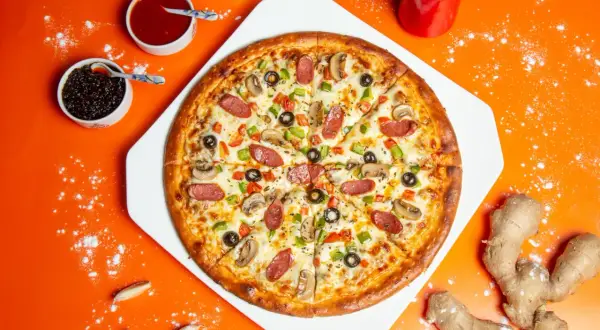 Pizza z pieczarkami, kiełbasą i warzywami na pomarańczowym tle, obok sosów i imbiru.