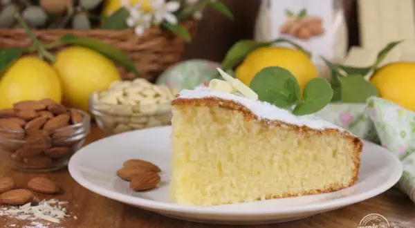 Una fetta di soffice torta caprese al limone, guarnita con zucchero a velo e foglioline di menta, circondata da limoni freschi e mandorle.