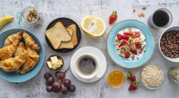 Un tavolo pieno di idee su cosa mangiare a colazione: croissant, toast, yogurt con frutta, caffè, uva e cereali.