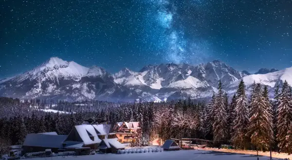 Zimowe Tatry nocą, Droga Mleczna nad ośnieżonymi szczytami i lasem. W dole przytulne górskie domki, idealne miejsce, gdzie w góry zima zaprasza.
