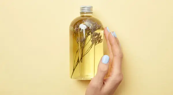 Eine Hand mit hellblauen Nägeln berührt eine Flasche mit gelbem Öl und getrockneten Blumen. Perfekt für alle, die Duschgel selber machen wollen.