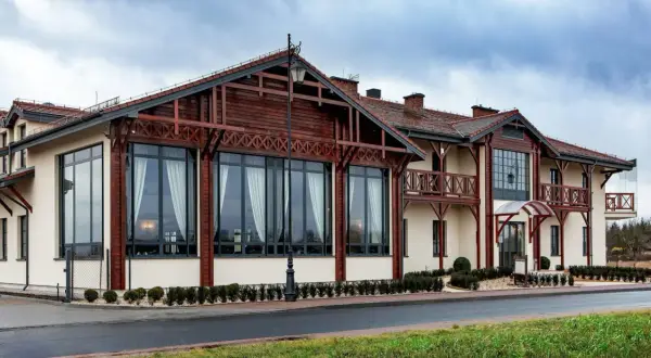 Nowoczesny budynek z drewnianymi elementami architektonicznymi, przypominający początek odyseja ciechocinek. Duże okna, balkony i zadbane otoczenie.