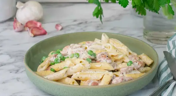 En krämig kyckling pasta med ärtor i en grön skål, med vitlök och persilja i bakgrunden.