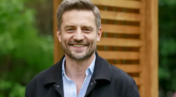 Uśmiechnięty Marcin Rogacewicz, znany z wielu filmów, pozuje na tle drewnianej pergoli i zieleni.