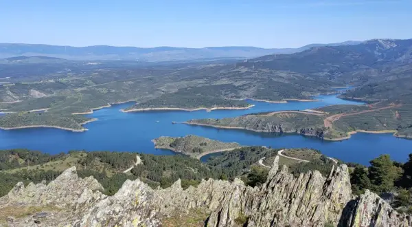 Vista panorámica del embalse de Puentes Viejas, con sus aguas azules serpenteando entre colinas verdes y montañas al fondo.
