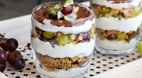 Dessert im Glas mit Schichten aus Creme, Keksbröseln, grünen und roten Trauben, garniert mit Kakaopulver.