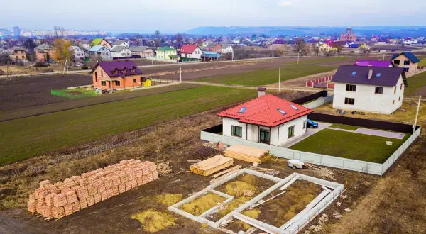 Budowa domku na działkę zrób to sam. Widać fundamenty, sterty cegieł i drewno budowlane. W tle ukończone domy.