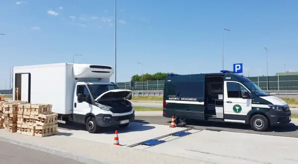 Kontrola drogowa: biała ciężarówka z otwartą maską i stosy palet obok, obok zielony bus Inspekcji Drogowej. Sprawdzana jest ładowność w dowodzie rejestracyjnym.