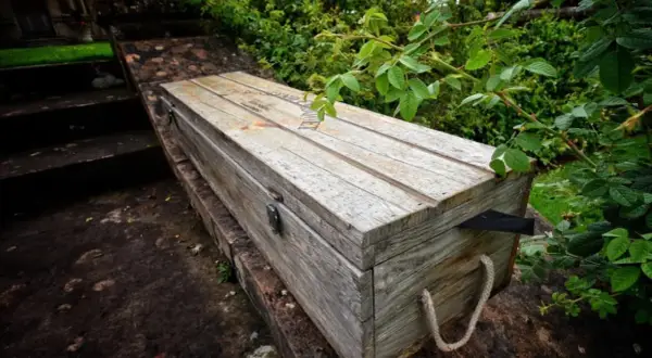 Drewniana skrzynia ogrodowa z linowymi uchwytami, na kamiennym murku, otoczona zielenią.