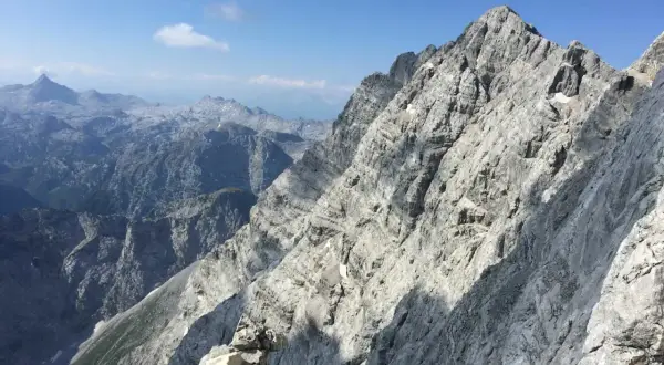 Steile Felsformationen der Watzmann Ostwand im Sonnenschein, mit tiefen Tälern und weiteren Berggipfeln in der Ferne.