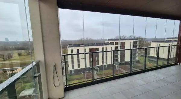 Zabudowa balkonu w bloku z widokiem na nowoczesne osiedle. Szklane panele tworzą przestronną, przeszkloną loggię.