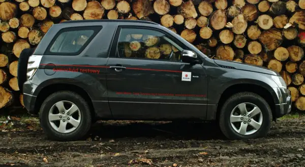 Suzuki Grand Vitara wymiary: ciemnoszary SUV testowy na tle stosu drewna.