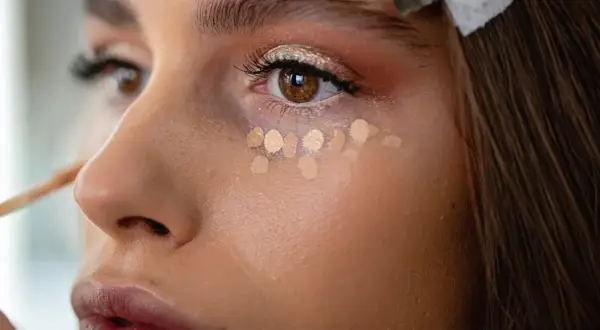 Nahaufnahme einer Frau, die Concealer unter den Augen aufträgt. Mehrere helle Tupfer bilden eine Reihe, bereit zum Verblenden.