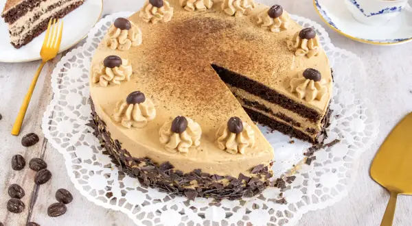 Eine köstliche Mokka Torte, verziert mit Kaffeebohnen und Schokostückchen, wartet auf Genießer. Daneben ein Stück Torte und eine Tasse Kaffee.