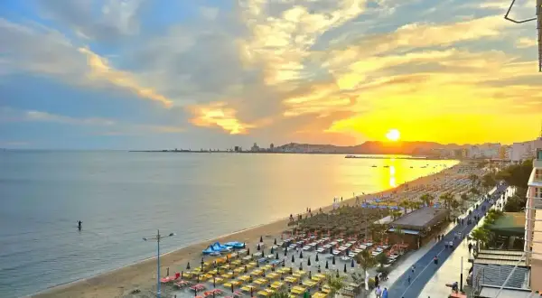 Zachód słońca nad Durrës plaża. Rzędy leżaków i parasoli czekają na plażowiczów, a w oddali widać panoramę miasta.
