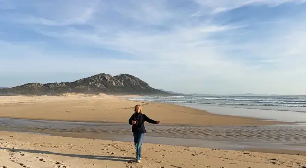 Mujer corre en la playa de Muros, Galicia, con montañas al fondo y el mar azul.