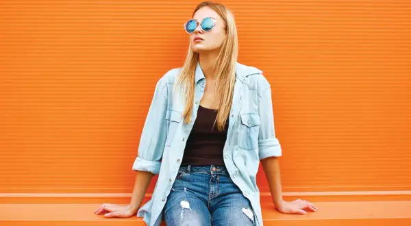 Blondynka w okularach i jeansach, z rozpiętą błękitną koszulą. Idealny przykład, jak nosić koszulę do jeansów na luzie.