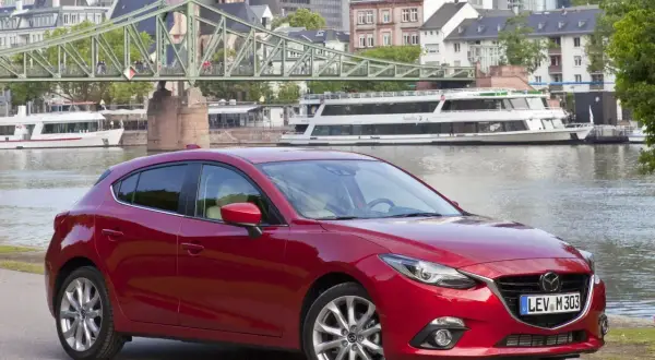 Czerwony sedan Mazda 3 na tle miasta z mostem i rzeką.