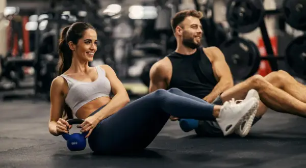Couple working out in a gym, doing Russian twists with a kettlebell. This is part of how to train for an Ironman.