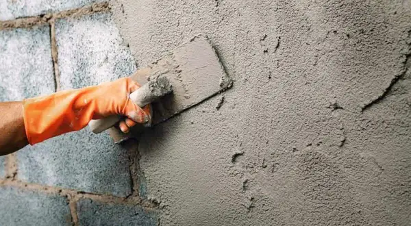 Mano con guante naranja aplica enfoscado rugoso a pared de bloques de cemento con llana.