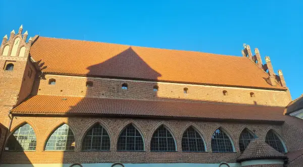 Zamek kapituły warmińskiej w Fromborku, ceglana budowla z pomarańczowym dachem i łukowymi oknami, pod błękitnym niebem.