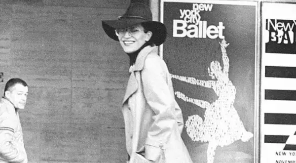 Elsa Peretti, con sombrero y gafas, sonríe frente a un cartel del New York City Ballet.