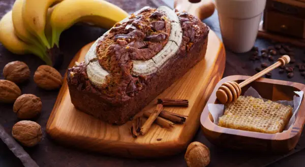Ein saftiges Bananenbrot mit Schokoladenstückchen und Nüssen, garniert mit einer Banane. Daneben Kaffee, Honigwabe und Zimtstangen – ein Genuss, der auch bananenbrot gesund macht.