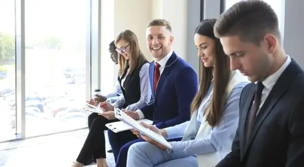 Giovani professionisti in attesa per un colloquio di gruppo, alcuni con appunti, uno sorride.