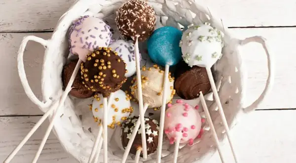 Kolorowe cake pops w białym koszyku, udekorowane posypkami i kuleczkami.