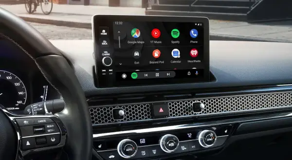 Wnętrze samochodu z ekranem Android Auto, na którym widać ikony Google Maps, Spotify i innych aplikacji. W tle dyskretnie widać Yanosik.