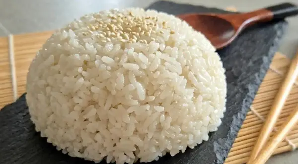 Ein Berg aus weißem Reis, bestreut mit Sesam, auf einem schwarzen Stein. Daneben liegen Stäbchen und ein Holzlöffel. Das ist die Basis, was bedeutet Sushi.
