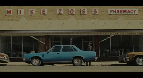 Niebieski samochód przed apteką "MIKE ZOSS PHARMACY". Obraz w stylu **false color**, z lekko zmienionymi barwami, tworzy nostalgiczny klimat.