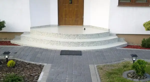 Nowe schody z żywicy na balkon, z kamyczkami, prowadzące do drewnianych drzwi. Wokół kostka brukowa i roślinność.