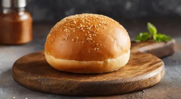 Ein goldbrauner Burger-Bun mit Sesam bestreut, liegt auf einem rustikalen Holzbrett.
