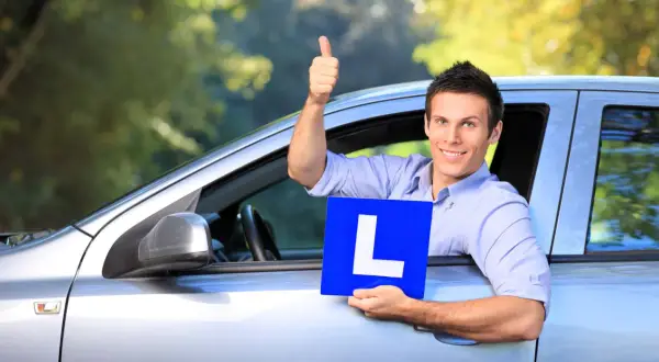 Hombre sonriente en coche con cartel "L", pulgar arriba. Pregunta: ¿cuanto tiempo hay que llevar la L?