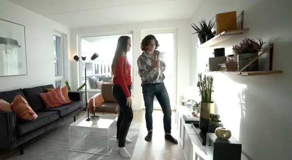 Två personer i ett ljust vardagsrum med stilren inredning. En mörk soffa med kuddar, ett vitt soffbord och en golvlampa. Hyllor med växter och dekorationer.