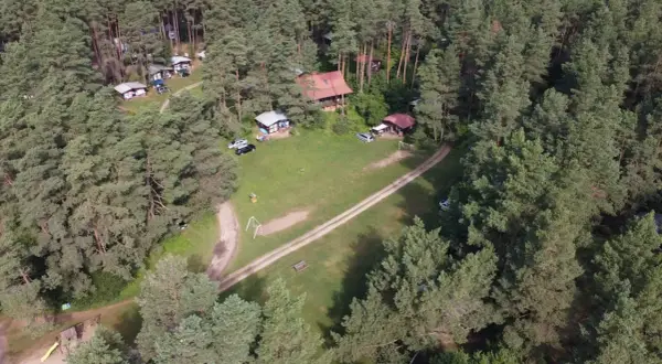 Malowniczy widok na stanicę wodną Zgon otoczoną lasem sosnowym. Widać domki, samochody i plac zabaw.