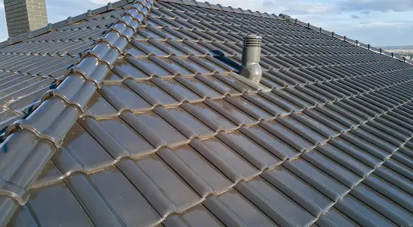 Szary dach z dachówki ceramicznej, z widocznym kominkiem wentylacyjnym i kominem. Rozmieszczenie kominków wentylacyjnych na dachu zapewnia prawidłową cyrkulację powietrza.