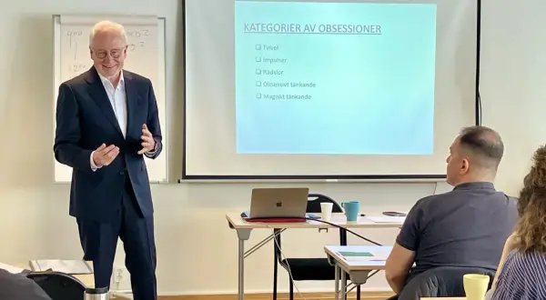 Föreläsare presenterar "Kategorier av obsessioner" som steg 1 i en utbildning. Publiken lyssnar uppmärksamt.