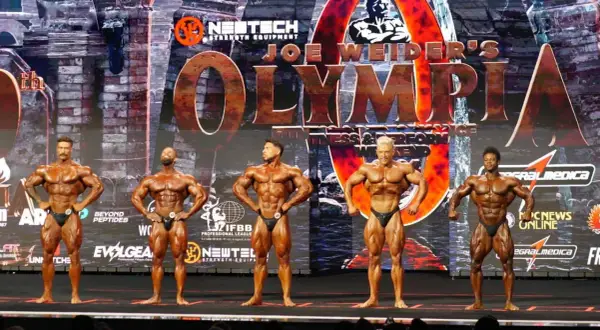 Fünf muskulöse Männer posieren auf der Bühne bei einem Bodybuilding-Wettkampf. Die "Joe Weider's Olympia" Bühne ist im Hintergrund zu sehen.