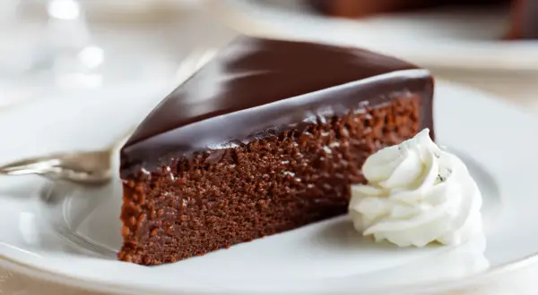 Ein Stück Sachertorte, wie im Sachertorte Rezept Original Lafer, mit glänzender Schokoladenglasur und einem Klecks Schlagobers auf einem weißen Teller.