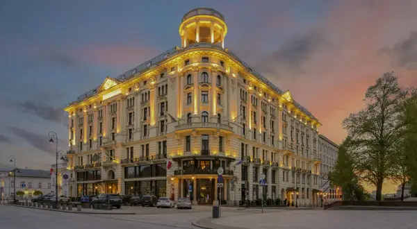 Elegancki budynek Mercure Warszawa Centrum oświetlony wieczorem, z charakterystyczną wieżyczką i ozdobną fasadą, na tle zachodzącego słońca.