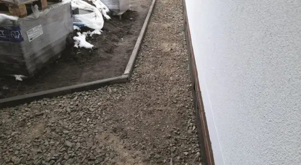 Ułożono kruszywo kamienne tłuczeń wzdłuż białego muru, obok palet z materiałami budowlanymi.